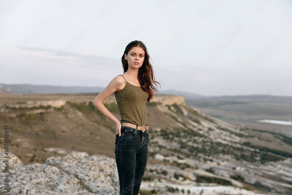 Woman in tank top and jeans stands triumphantly atop mountain, overlooking picturesque valley below Woman in tank top and jeans stands triumphantly atop mountain, overlooking picturesque valley below