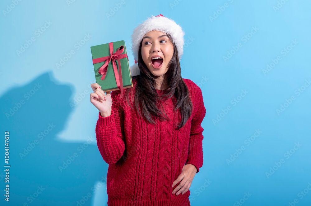 Obraz premium Asian woman, wearing a Santa hat and a red knitted sweater, looks surprised as she holds a green gift box tied with a red ribbon. She is in front of a light blue background