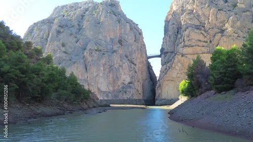 Drone flight over the river to the gorge Garganta del Chorro and Caminito del Rey, Spain. A bridge in a narrow gorge between huge high mountains, green forest on the river banks. 