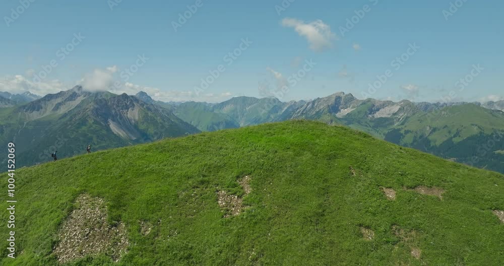 Two hikers trek a grassy hilltop with a vast mountain range in the background under a clear sky Two hikers trek a grassy hilltop with a vast mountain range in the background under a clear sky