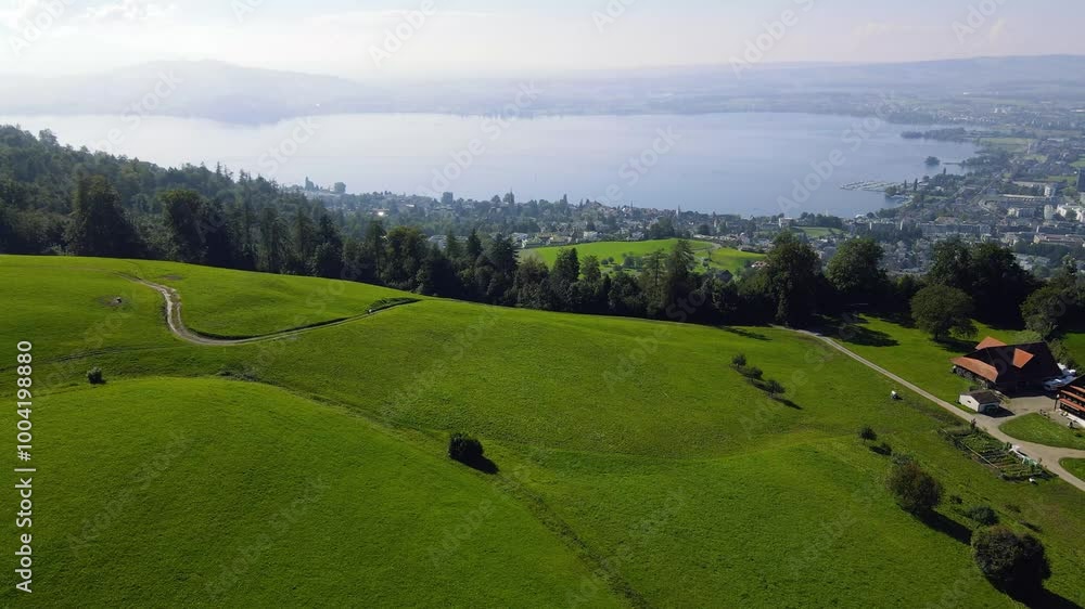 A horizontal panoramic shot of Alpine Swiss farms surrounded by green fields and overlooking the spectacular Lake Zug in a hazy atmosphere in Switzerland A horizontal panoramic shot of Alpine Swiss farms surrounded by green fields and overlooking the spectacular Lake Zug in a hazy atmosphere in Switzerland