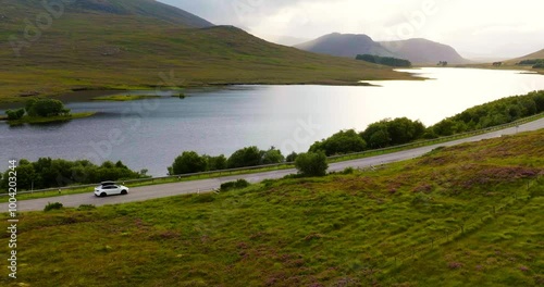 Aerial shot of white electro car driving with breathtaking Scottish landscape