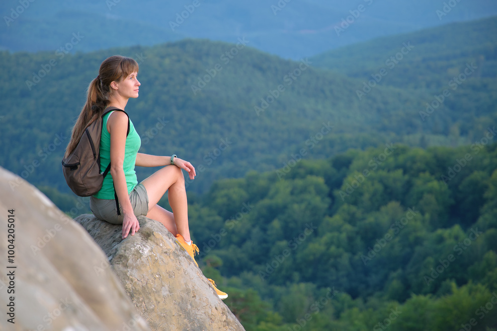 Naklejka premium Woman hiker seated alone on rocky mountain cliff enjoying view of evening nature on wilderness trail. Active way of life concept