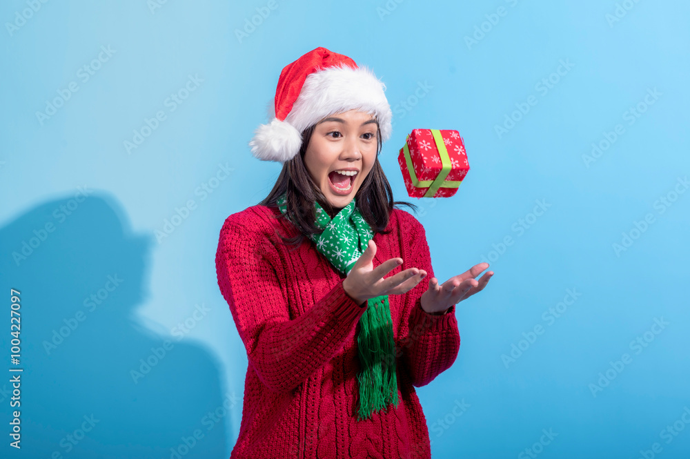 Obraz premium Asian woman in a red sweater, Santa hat, and green scarf is joyfully tossing a red gift box with snowflake patterns and a green ribbon in the air. She stands against a light blue background