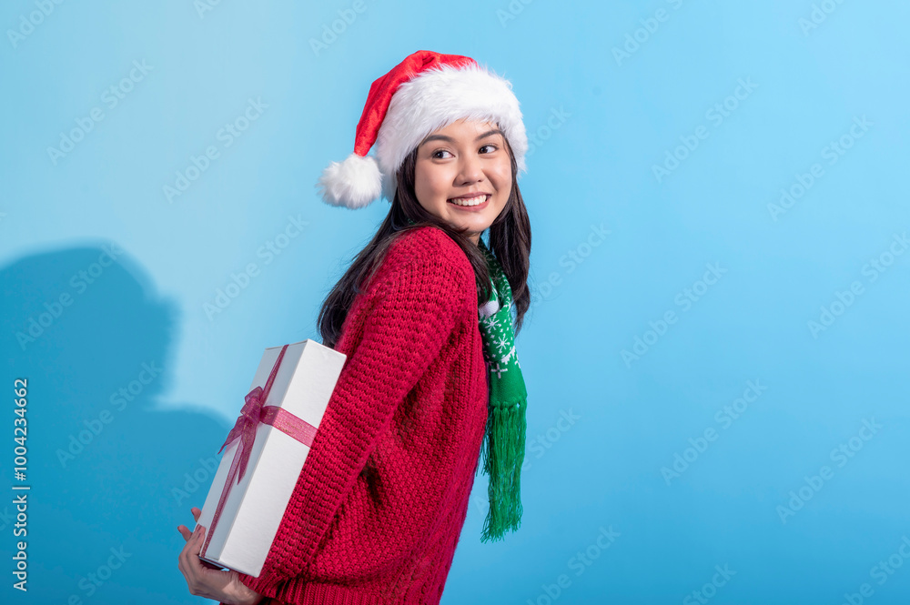 Obraz premium An Asian woman wearing a Santa hat, red sweater, and green scarf smiles playfully while holding a white gift box tied with a pink ribbon behind her back. She stands against a light blue background