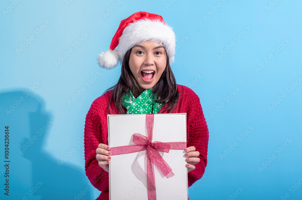 An Asian woman wearing a Santa hat, red sweater, and green scarf smiles warmly while holding a white gift box tied with a pink ribbon. She stands against a light blue background An Asian woman wearing a Santa hat, red sweater, and green scarf smiles warmly while holding a white gift box tied with a pink ribbon. She stands against a light blue background