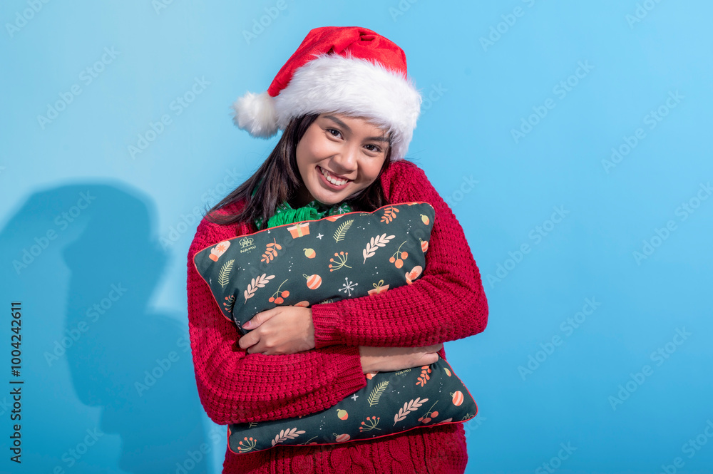 An Asian woman dressed in a red sweater and Santa hat smiles warmly as she hugs a Christmas-themed pillow decorated with holiday patterns. She stands against a light blue background An Asian woman dressed in a red sweater and Santa hat smiles warmly as she hugs a Christmas-themed pillow decorated with holiday patterns. She stands against a light blue background
