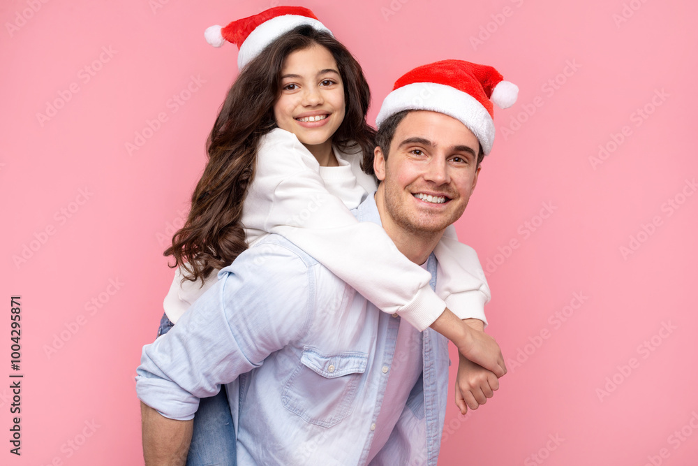Xmas mood. Excited father and daughter wearing Santa hats, dad piggybacking girl, having fun on pink background and smiling at camera Xmas mood. Excited father and daughter wearing Santa hats, dad piggybacking girl, having fun on pink background and smiling at camera