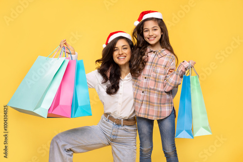 Young woman and her child girl both in Santa hats, holding shopping package bags isolated on yellow background. Xmas winter shopping concept photo