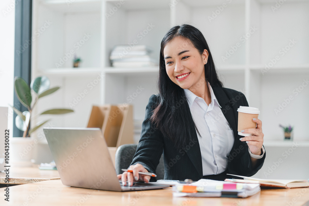professional woman in black suit smiles while working on laptop in modern office. She holds coffee cup, showcasing productive and positive work environment. professional woman in black suit smiles while working on laptop in modern office. She holds coffee cup, showcasing productive and positive work environment.