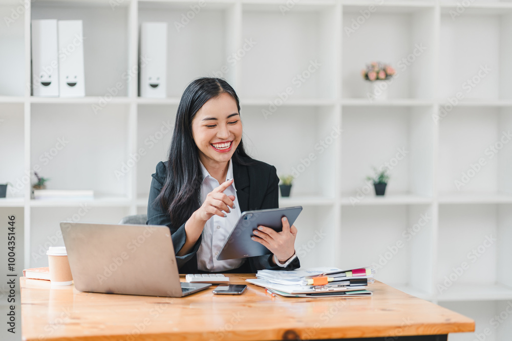 young woman in black suit smiles while using tablet at her desk, surrounded by office supplies and laptop. Her cheerful demeanor reflects productive work environment. young woman in black suit smiles while using tablet at her desk, surrounded by office supplies and laptop. Her cheerful demeanor reflects productive work environment.