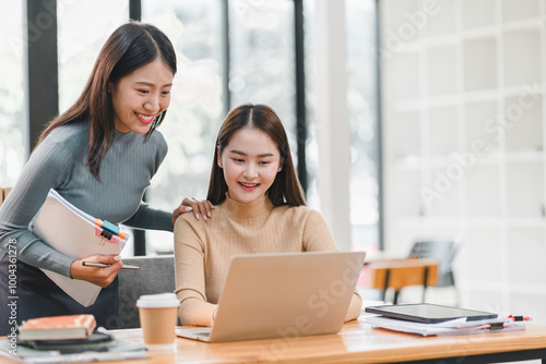 Two women collaborating in modern office, one assisting other with laptop. They appear engaged and focused, showcasing teamwork and productivity.