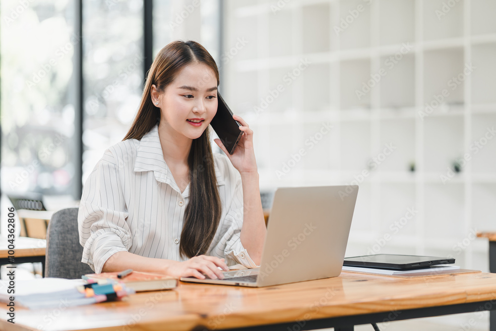 young woman is engaged in phone conversation while working on her laptop in modern office setting. She appears focused and productive, surrounded by bright and organized workspace. young woman is engaged in phone conversation while working on her laptop in modern office setting. She appears focused and productive, surrounded by bright and organized workspace.