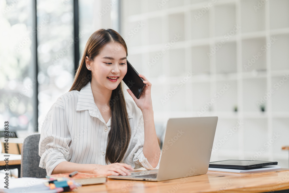young woman is engaged in phone conversation while working on her laptop in modern office setting. She appears focused and productive, showcasing blend of technology and communication. young woman is engaged in phone conversation while working on her laptop in modern office setting. She appears focused and productive, showcasing blend of technology and communication.