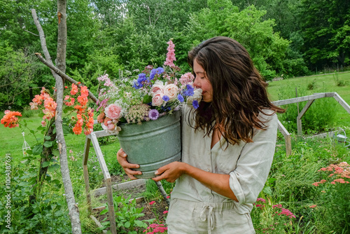 A woman in a light dress smiles as she holds a bucket brimming with colorful flowers in a lush garden. photo