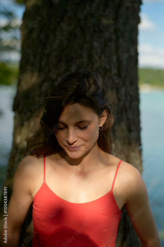 Serene woman in a red dress standing beside a tree with a lake in the background. Serene woman in a red dress standing beside a tree with a lake in the background.