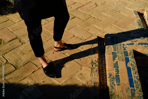 Sunlight casts the long shadow of a person walking on a patterned tile pavement. photo