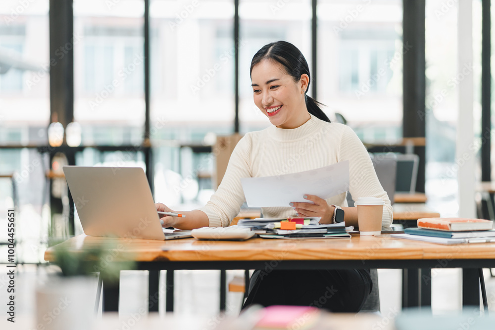 young woman is working at desk in modern office, smiling as she reviews documents on her laptop. bright environment enhances her focused and positive demeanor. young woman is working at desk in modern office, smiling as she reviews documents on her laptop. bright environment enhances her focused and positive demeanor.