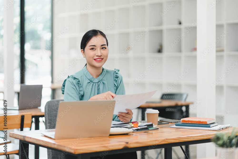 young woman in teal blouse sits at wooden desk, smiling while holding documents. modern office setting features laptop, notebooks, and coffee cup, creating productive atmosphere. young woman in teal blouse sits at wooden desk, smiling while holding documents. modern office setting features laptop, notebooks, and coffee cup, creating productive atmosphere.