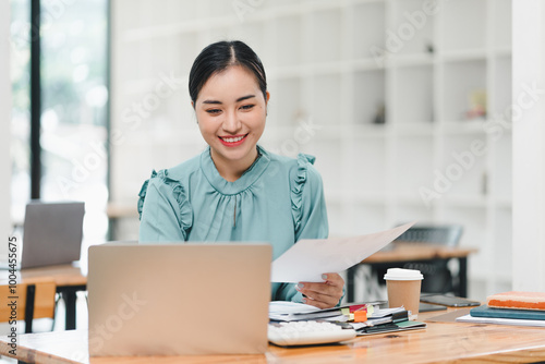 woman is smiling while working on laptop in modern office environment, holding documents in her hand. atmosphere is bright and productive, reflecting positive work experience.