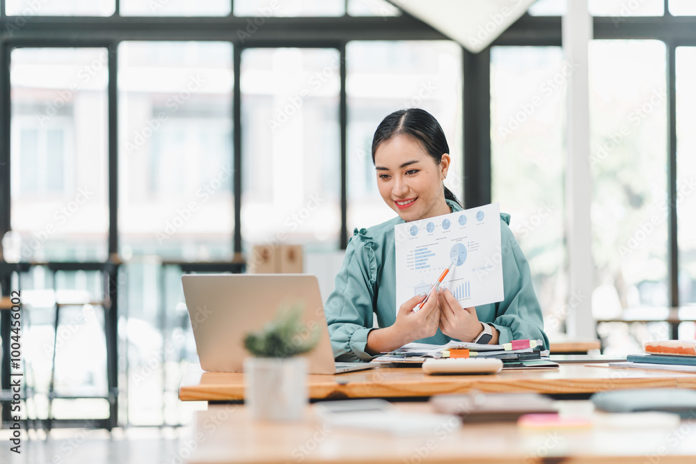 Fototapeta premium young woman is engaged in video conference, presenting data with chart in modern office setting. She appears focused and confident while using laptop and various documents on table.