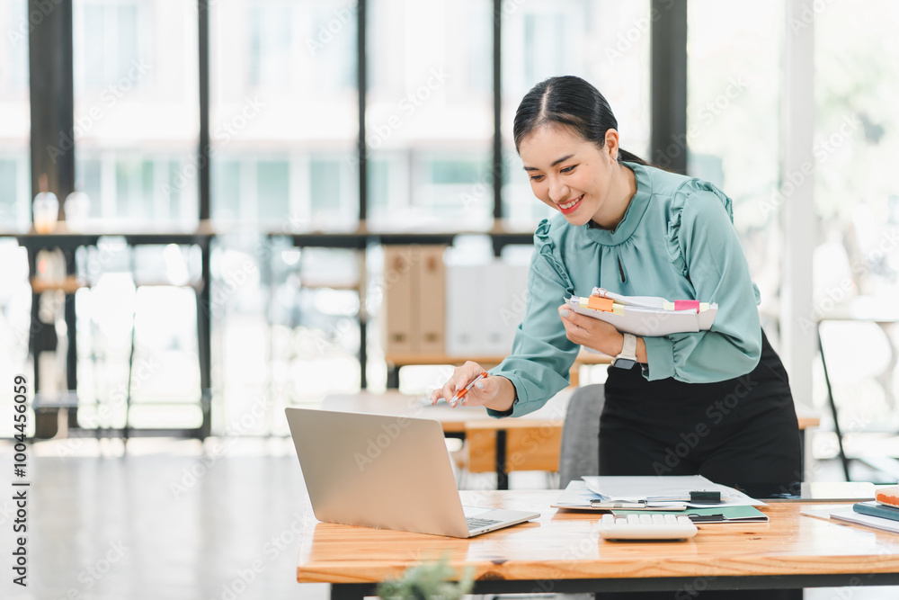 professional woman is smiling while working on laptop in modern office environment. She holds notebook and appears engaged in her tasks, showcasing positive and productive atmosphere. professional woman is smiling while working on laptop in modern office environment. She holds notebook and appears engaged in her tasks, showcasing positive and productive atmosphere.
