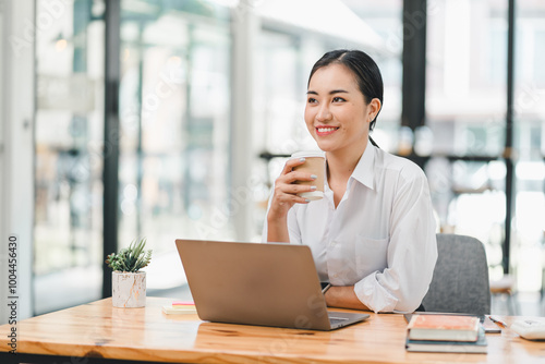 young woman is sitting at wooden table, smiling while holding coffee cup. She is working on laptop in bright, modern office space, surrounded by books and small plant.