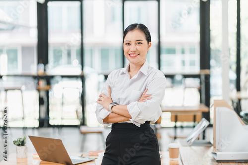 confident woman stands with her arms crossed in modern office setting, showcasing professionalism and poise. bright workspace features laptop and welcoming atmosphere.