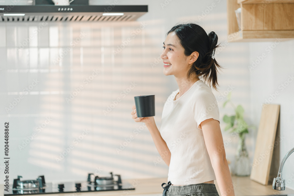 woman stands in modern kitchen, holding black cup and smiling. sunlight casts soft shadows, creating warm and inviting atmosphere. woman stands in modern kitchen, holding black cup and smiling. sunlight casts soft shadows, creating warm and inviting atmosphere.