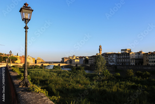 Lamp Post and Cityscape View Along Arno River in Florence.A serene evening shot of Florence, featuring a classic street lamp, the Arno River, and the city's historic buildings in the soft light.