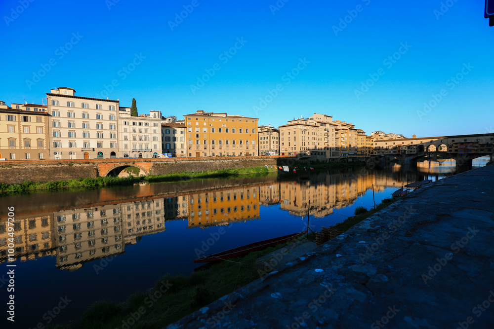 Fototapeta premium Reflections of Historic Florence along the Arno River The bright facades of buildings in Florence reflect perfectly on the calm waters of the Arno River, with the Ponte Vecchio in the distance.