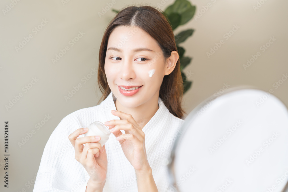 Facial beauty skin care, smile of pretty asian young woman in bathrobe looking at mirror, hand applying moisturizer lotion on her face, holding jar of skin cream before makeup cosmetic routine at home