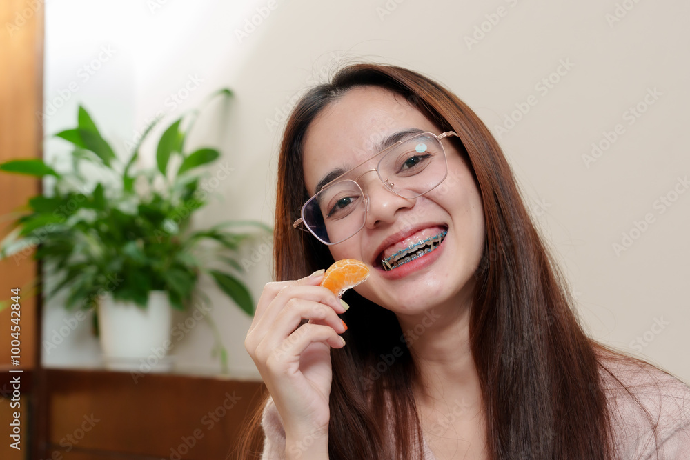 Asian woman with glasses smiling while holding a peeled orange slice in a cozy bedroom setting. Bright and cheerful mood reflects happiness and relaxation in a warm, indoor environment. Asian woman with glasses smiling while holding a peeled orange slice in a cozy bedroom setting. Bright and cheerful mood reflects happiness and relaxation in a warm, indoor environment.