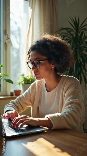 A woman is sitting at a table with a laptop in front of her. She is wearing glasses and she is focused on her work. The room has a window with a potted plant in front of it