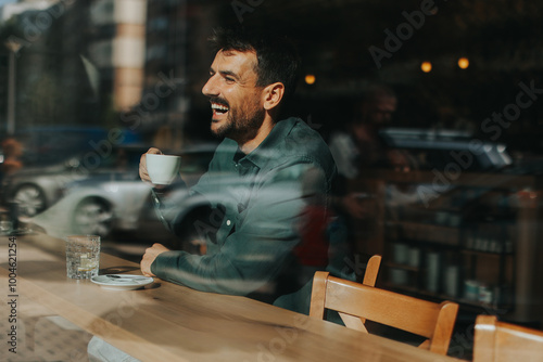 Laughing man enjoys a warm cup of coffee while seated at a sunlit cafe, basking in a cheerful afternoon atmosphere with city views