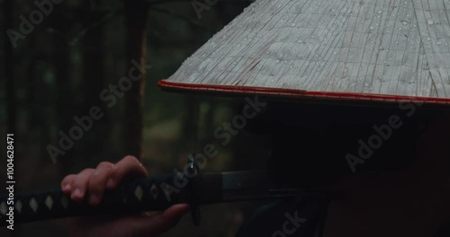 A man with a katana in the forest. A warrior in a hat unsheathes a katana in the forest, raindrops falling on his hat. The scene is a combination of nature and martial arts in a cinematic atmosphere. photo