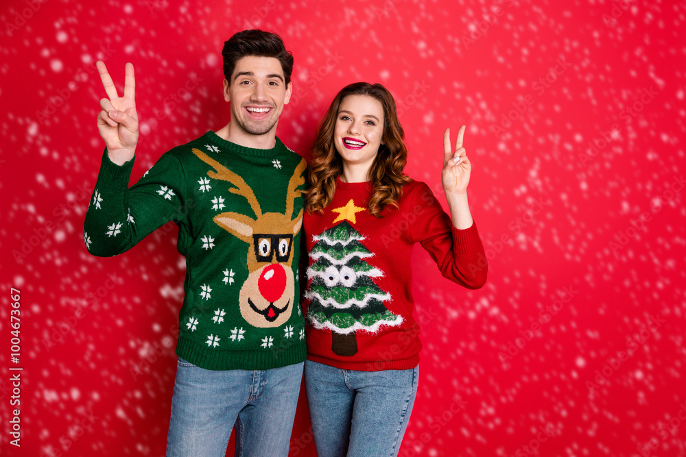 Portrait of cheerful two hipsters make v-signs wear deer christmas tree design patterns pullover jumper isolated over red color background Portrait of cheerful two hipsters make v-signs wear deer christmas tree design patterns pullover jumper isolated over red color background