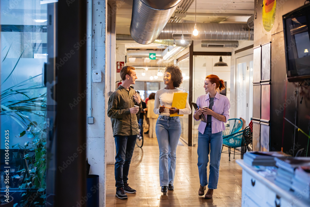 Obraz premium Three multiracial coworkers walking through coworking office. Group of young coworkers walking and talking in a brightly lit office hallway, carrying documents and tablets.