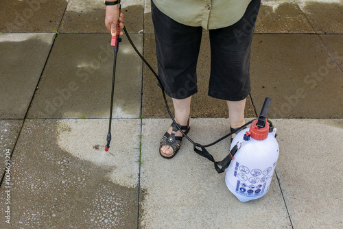 Woman spraying gray concrete patio paving slabs with liquid weed killer from pressurized plastic tank and handheld nozzle photo