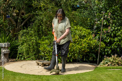 Woman in black trouser pants and wellie boots using electric strimmer weed whacker to trim grass lawn edge photo