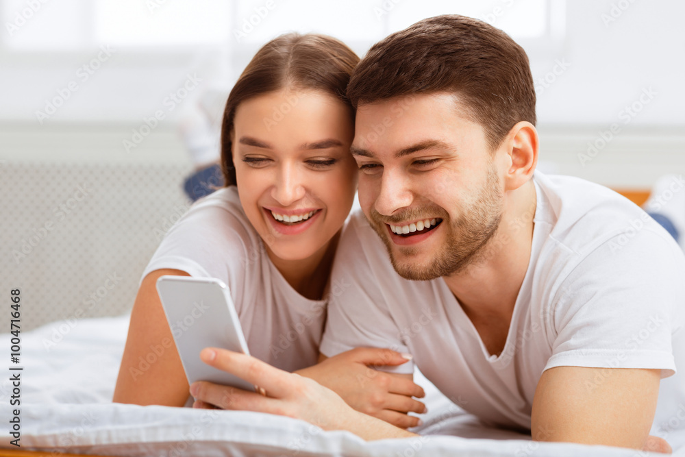Fototapeta premium Mobile App. Cheerful Couple Using Smartphone Lying In Bed At Home In The Morning. Selective Focus