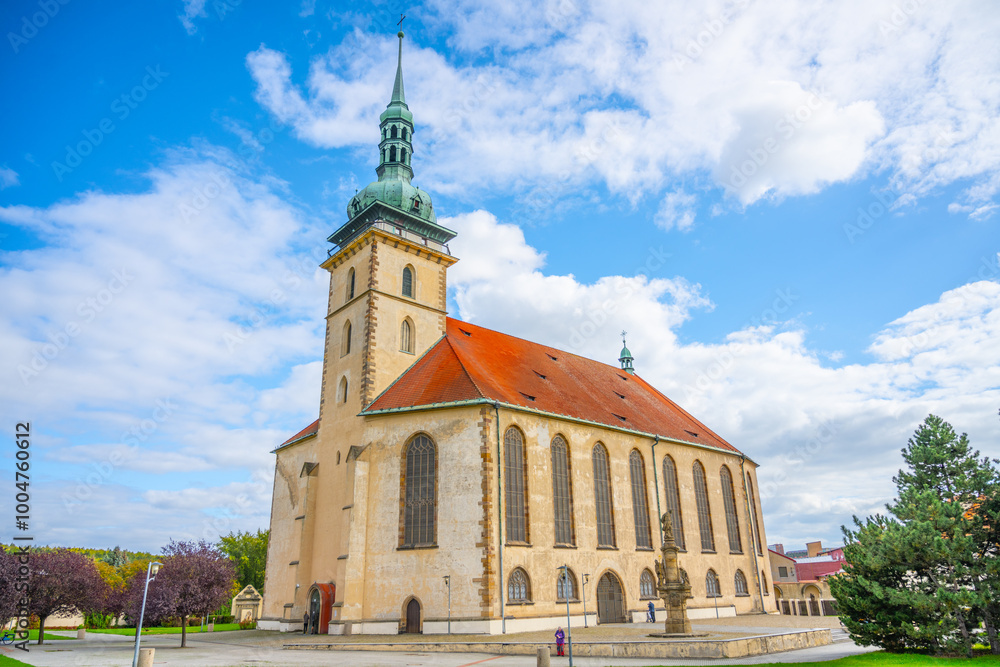Naklejka premium The Church of the Assumption of the Virgin Mary stands prominently in Most, Czechia, showcasing late Gothic architecture against a vibrant sky, surrounded by lush greenery.