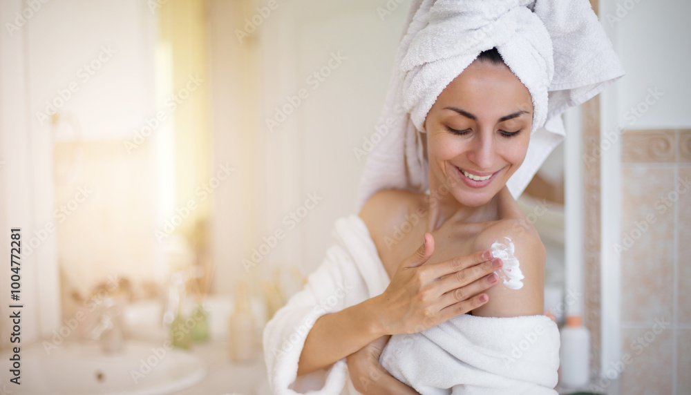 Beautiful smiling woman with a towel wrapped around her head, applying lotion or cream to her shoulder in a well-lit bathroom. She wears a white bathrobe, radiating relaxation and self-care. Beautiful smiling woman with a towel wrapped around her head, applying lotion or cream to her shoulder in a well-lit bathroom. She wears a white bathrobe, radiating relaxation and self-care.