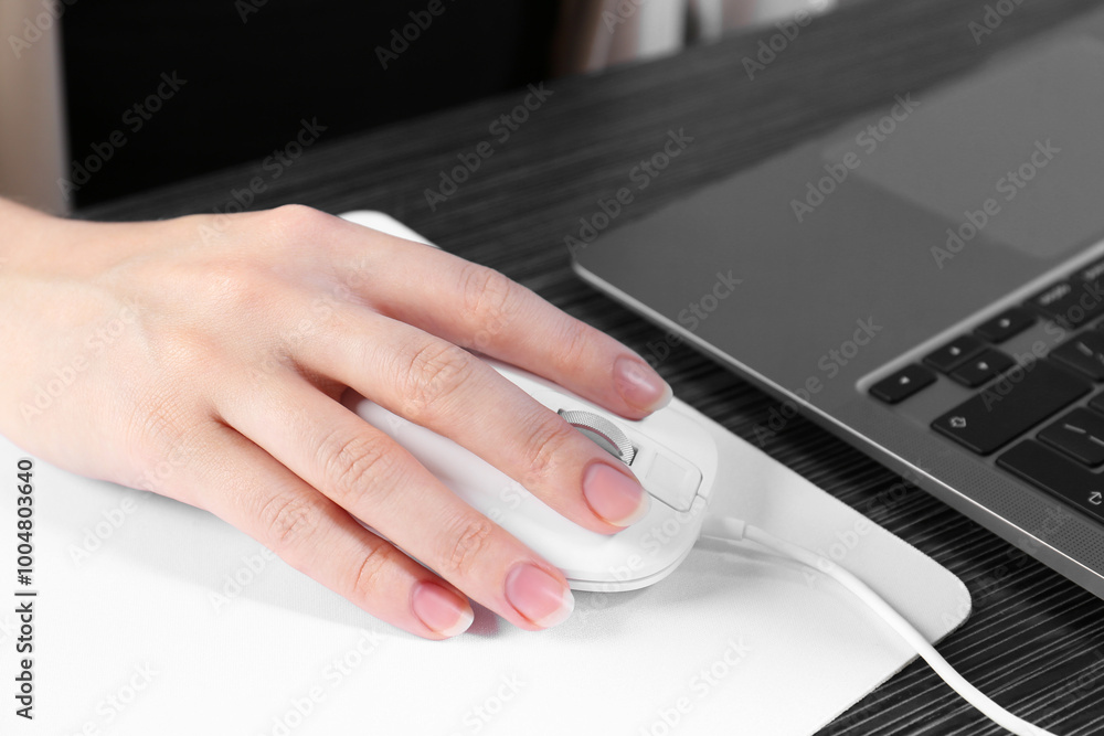 Fototapeta premium Woman using computer mouse while working with laptop at black wooden table, closeup