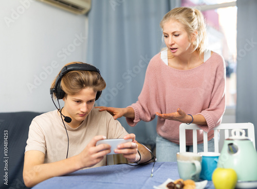 Focused teenager carried away with smartphone not paying attention to his worried mother talking to him photo