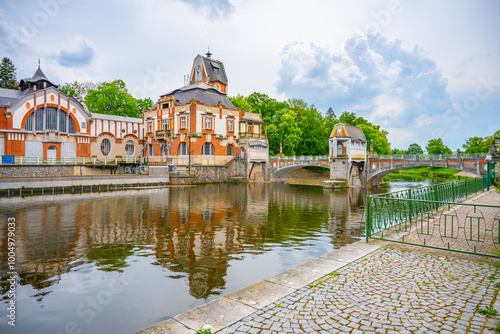 The historic Art Nouveau Hucak hydroelectric power station stands beside a tranquil river in Hradec Kralove, Czechia, showcasing intricate design and surrounded by lush greenery under a cloudy sky. photo