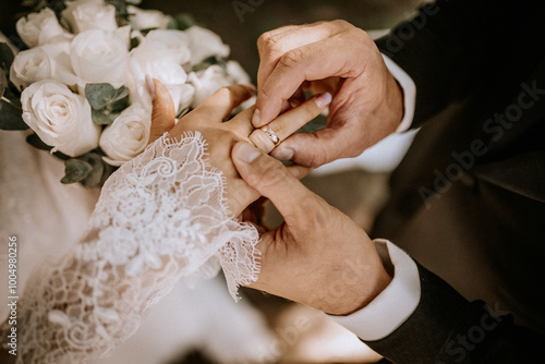 A close-up of a groom placing a wedding ring on the bride’s finger during the ceremony, with her lace sleeve and white rose bouquet visible.