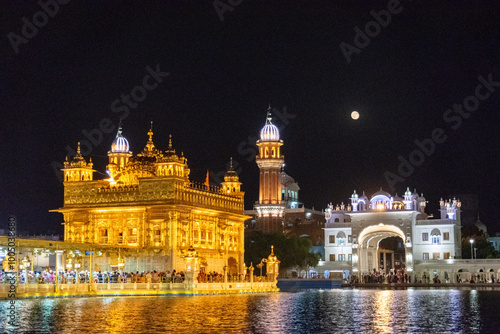 The golden temple harmandir sahib is shining on amrit sarovar lake at night with full moon in photo