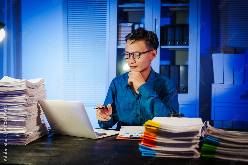 Asian businessman works late into the night, focused on a stack of papers on his black wooden desk. The office is dimly lit with a blue glow, reflecting the busy atmosphere of overtime. Asian businessman works late into the night, focused on a stack of papers on his black wooden desk. The office is dimly lit with a blue glow, reflecting the busy atmosphere of overtime.