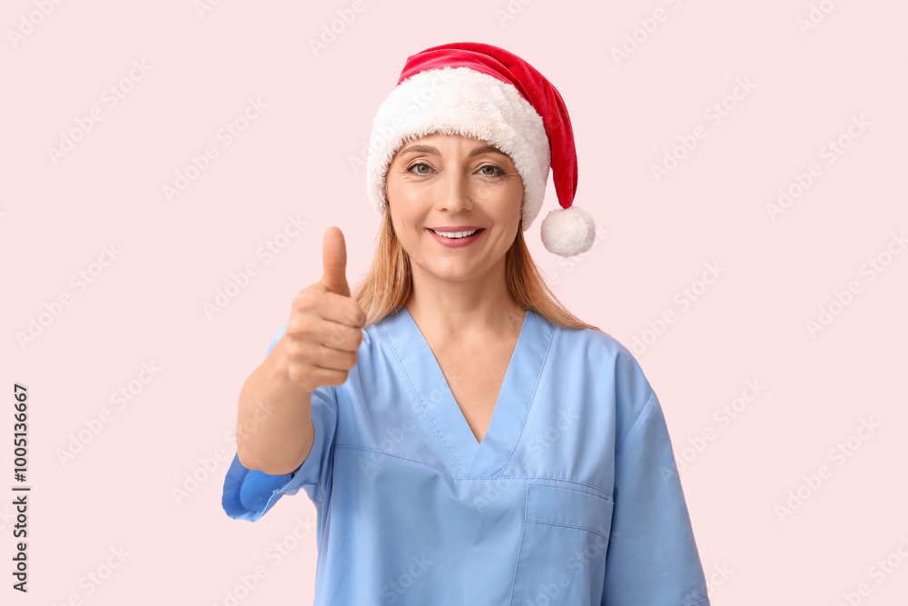 Mature female doctor in Santa hat showing thumb-up gesture on pink background Mature female doctor in Santa hat showing thumb-up gesture on pink background
